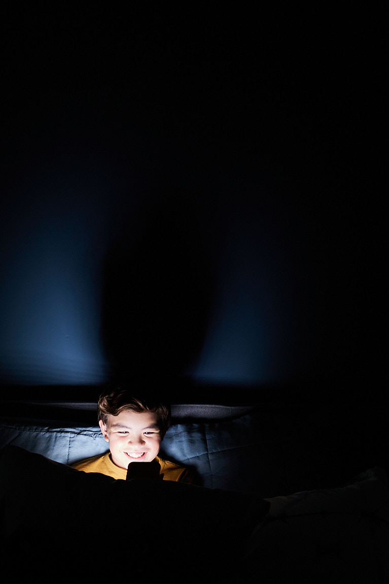A young kid looks at a phone in bed. 